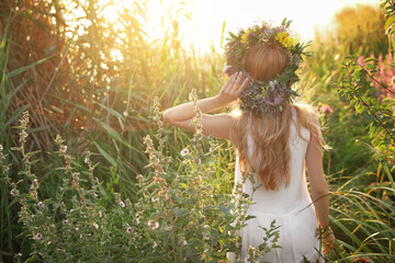 Young woman wearing wreath made of beautiful flowers outdoors on sunny day, back view