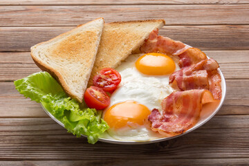 White plate of fried eggs with bacon, toasts and vegetables on wooden table, top view.