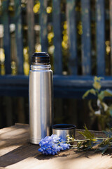 Thermos with tea and a purple flower on the background of nature and a fence. Stop in the journey. Wood texture