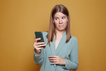 Dissatisfied lovely woman looks with disgust, aversion at cellular, poses over yellow background, receives call from boyfriend, holds mobile phone on distance, ignores communication, dislike something