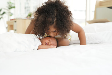 Portrait of African newborn baby sleeping on a white bed. His little sister kissing infant boy with love. New siblings relationship in bedroom at home