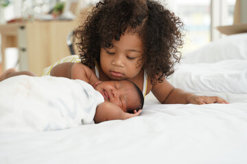 Portrait of African newborn baby sleeping on a white bed. His little sister kissing infant boy with love. New siblings relationship in bedroom at home