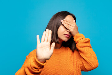 hispanic woman covering face with hand and putting other hand up front to stop camera, refusing...