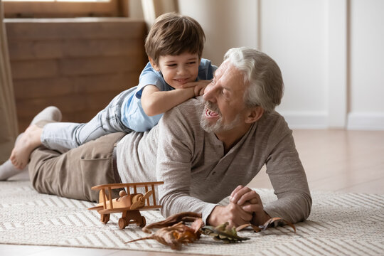 Overjoyed Mature Grandfather Playing With Adorable Grandson At Home, Older Man Lying On Warm Floor With Cute Little Boy Grandchild On Back, Family Spending Leisure Time Together, Having Fun