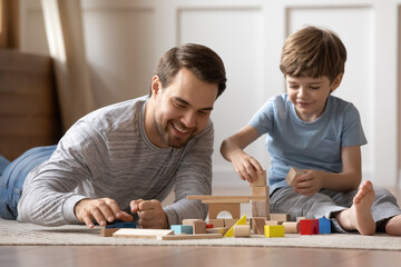 Close up happy loving father playing with adorable son at home, lying on warm floor, smiling dad and little boy kid constructing with wooden colorful blocks, enjoying leisure time in living room
