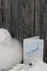 Winter knitted hat with fur and gloves. White. Nearby is a greeting card, a medical mask and an antiseptic. They lie on black pine boards.