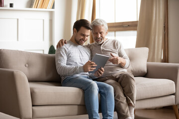 Happy mature father and adult son using tablet at home together, smiling older grandfather and grandson looking at device screen, sitting on couch, young man teaching senior dad to use computer