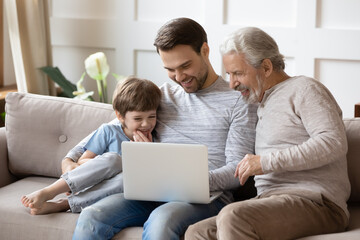 Smiling little boy with father and mature grandfather using laptop, sitting on couch, happy three...