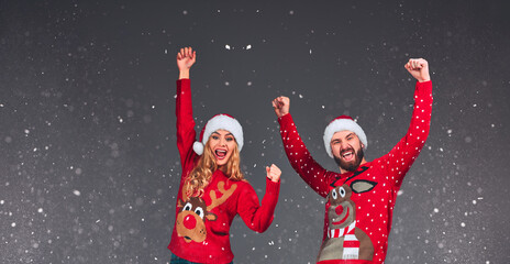 Festive couple in Santa hats smiling with arms raised on grey background.