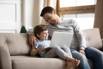 Happy father and little boy using laptop at home together, browsing apps, playing game, enjoying...