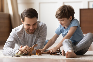 Close up happy loving father and adorable son playing with toy dinosaurs, sitting on warm floor at...