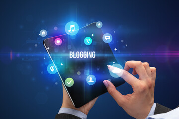 Businessman holding a foldable smartphone with BLOGGING inscription, social media concept