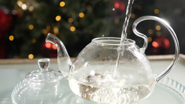 Christmas time and New Year atmosphere. Brewing tea with boiling water with New Year fir-tree in background. Making tea in slow motion. Adding water to jasmine tea flower in glass teapot. Full hd