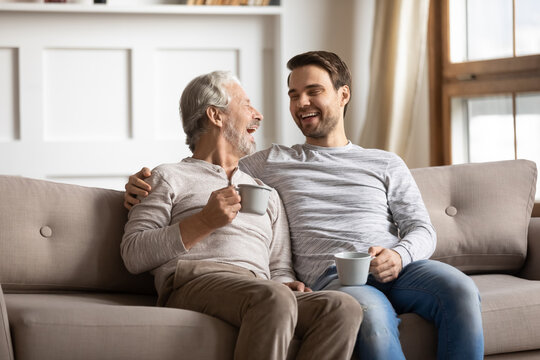 Overjoyed mature father and adult son drinking tea together, chatting, enjoying pleasant conversation, happy older grandfather and grandson laughing at funny joke, sitting on couch at home