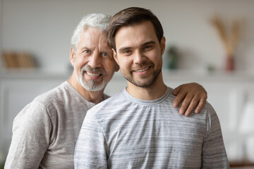Head shot portrait smiling mature father and adult son hugging, standing, posing for photo at home,...