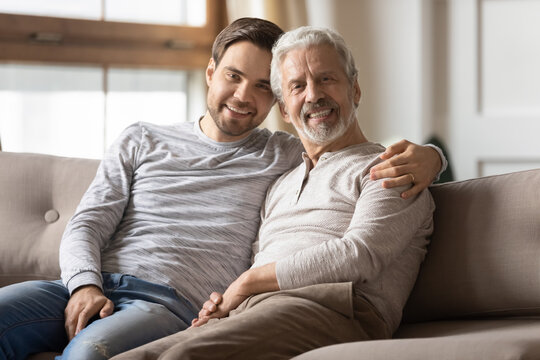 Portrait smiling young man hugging mature father, sitting on cozy couch at home, happy senior grandfather with grandson looking at camera, excited adult son with senior dad posing for picture