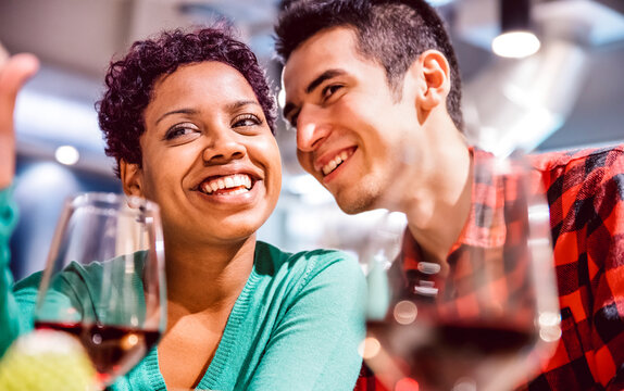 Happy Mixed Race Couple Of Lovers Drinking Red Wine At Fancy Winery Bar - Handsome Man Whispers Soft Kisses In Beautiful Woman Ear - Relationship Concept With Boyfriend And Girlfriend On Vivid Filter