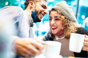 Happy couple with face mask having fun together at bar cafeteria - New normal lifestyle concept with young people talking and drinking american coffee - Bright azure filter with focus on woman eye
