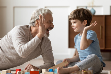 Fototapeta premium Close up happy mature grandfather and grandson having fun, playing with toys on floor together, older man and little boy giving high five, enjoying leisure time, constructing with wooden blocks