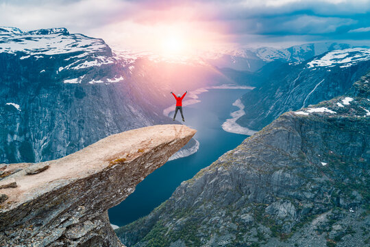 Norway, A woman Jumping on the mountains cliff edge of Trolltunga throning over Ringedalsvatnet  watching the sunset and snowy Norwegian mountains near Odda, Rogaland, Norway