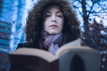 Young latin woman reads a book in the park during the winter season, wearing a hooded coat and a shawl