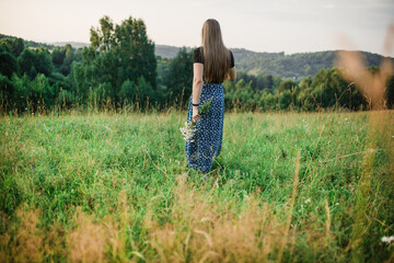 The girl walks across the field. Portrait of  woman from behind in a long dress walking along the road in a green field. slender young woman with long hair stands in a field with a bouquet of daisies