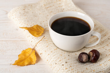 Large white mug of hot steaming coffee on a beige knitted scarf and two yellow fall leaves over white wood rustic surface. Cozy autumn mood concept.