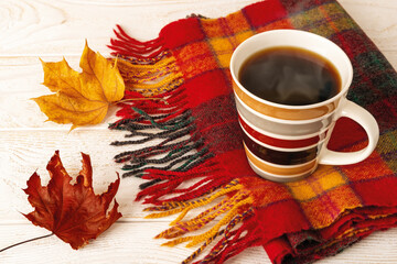 Striped mug of hot steaming coffee on a fringed wool check scarf and two autumn leaves over white...
