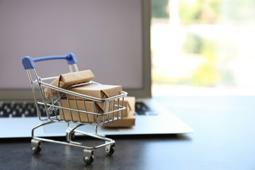 Internet shopping. Small cart with boxes near modern laptop on table indoors, space for text