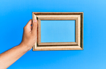 Hand of young hispanic man holding vintage empty frame over isolated blue background.