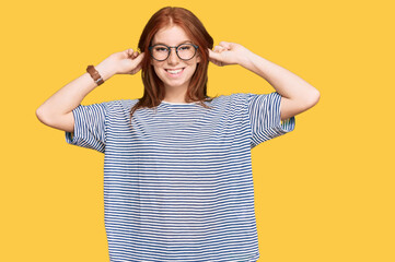Young read head woman wearing casual clothes and glasses smiling pulling ears with fingers, funny...