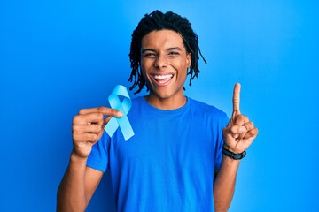 Young african american man holding blue ribbon smiling with an idea or question pointing finger with happy face, number one