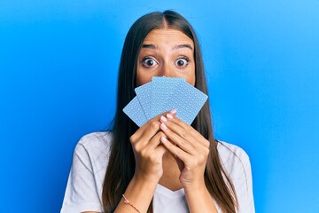 Young hispanic woman playing gambling poker covering face with cards celebrating crazy and amazed for success with open eyes screaming excited.