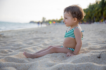 cute toddler in a swimsuit sits on a sandy tropical beach of the warm sea in the sunshine