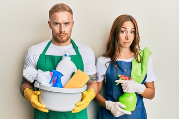 Young couple of girlfriend and boyfriend wearing apron holding products and cleaning spray skeptic and nervous, frowning upset because of problem. negative person.