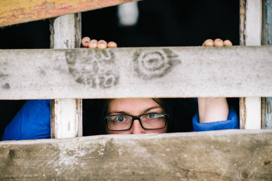 Locked up guilty girl in glasses emotional portrait. Woman slavery. Werid strange unusual person looking through slit in boarded up window in old wooden abandoned ghost house. Female eyes expression.