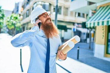 Young redhead architect man with long beard wearing hardhat smiling happy. Having conversation talking on the smartphone holding blueprints at street of city.