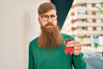 Young irish man with serious expression holding red hiv ribbon leaning on the wall at the city.