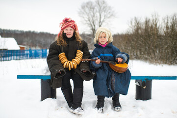 Two beautiful village girls sit on the bench with balalaika and barankas in winter