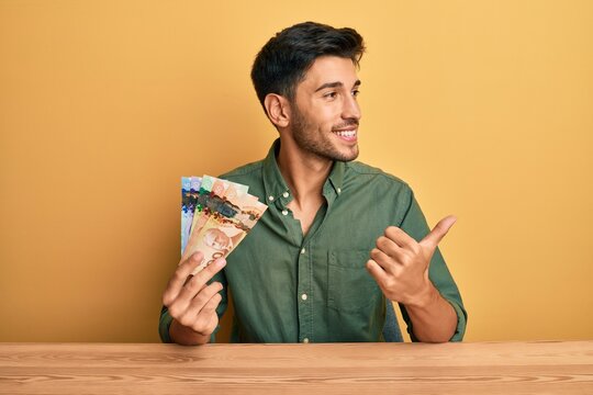 Young handsome man holding canadian dollars smiling with happy face looking and pointing to the side with thumb up.
