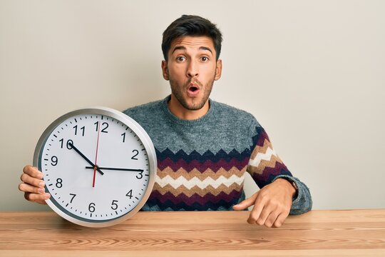 Young handsome man holding big clock scared and amazed with open mouth for surprise, disbelief face