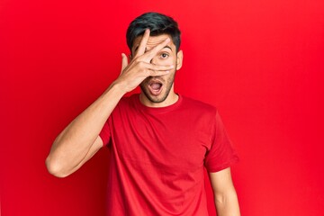 Young handsome man wearing casual tshirt over red background peeking in shock covering face and eyes with hand, looking through fingers afraid