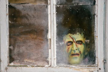 A child in a terrible monster mask peeks out of an old dirty window in winter