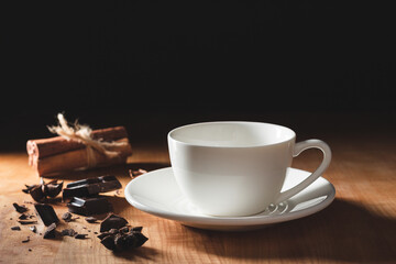 Empty white coffee cup or cappuccino mug with chopped chocolate, cinnamon stick and star anise on wooden table with dark background
