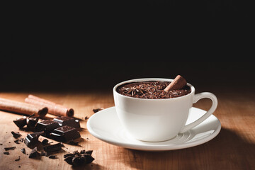 Hot chocolate drink in white cup, broken chocolate, cinnamon stick and star anise on a wooden table with dark background