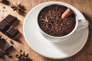 Hot chocolate drink in white cup, broken chocolate, cinnamon stick and star anise on a wooden table