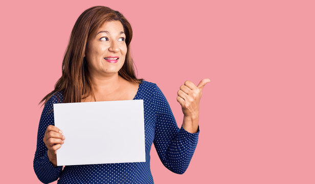 Middle age latin woman holding paper banner with blank space pointing thumb up to the side smiling happy with open mouth