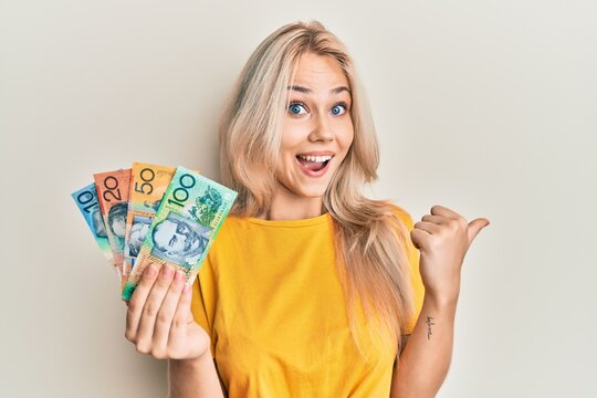 Beautiful caucasian blonde girl holding australian dollars pointing thumb up to the side smiling happy with open mouth