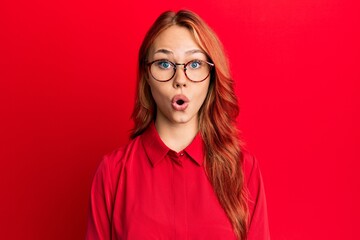Young beautiful redhead woman wearing casual clothes and glasses over red background afraid and shocked with surprise expression, fear and excited face.