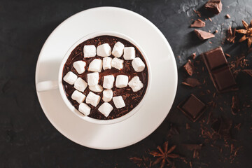Hot chocolate drink in a white cup with marshmallow on textured dark background, top view